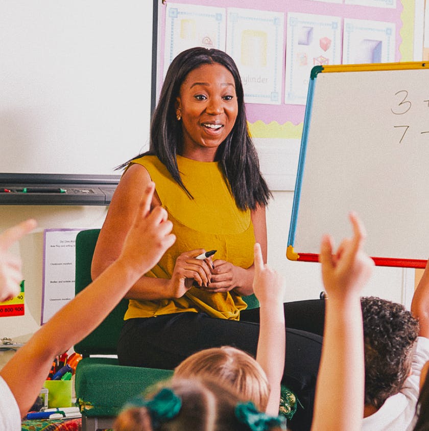 A teacher stands in front of a whiteboard while children have their hands raised
