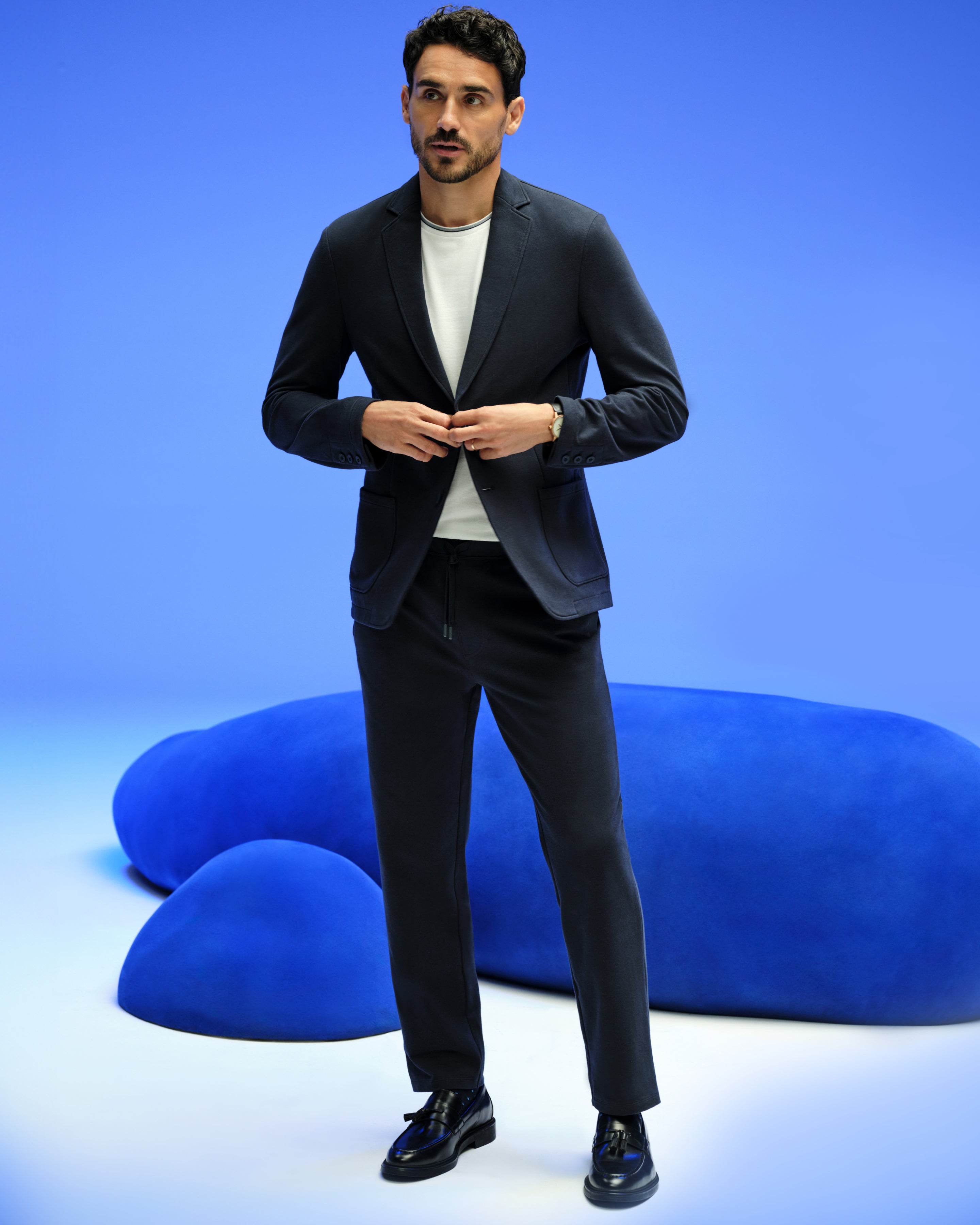 A man wearing slacks and a crew neck t-shirt, he is standing in a blue room and is buttoning up his blazer.