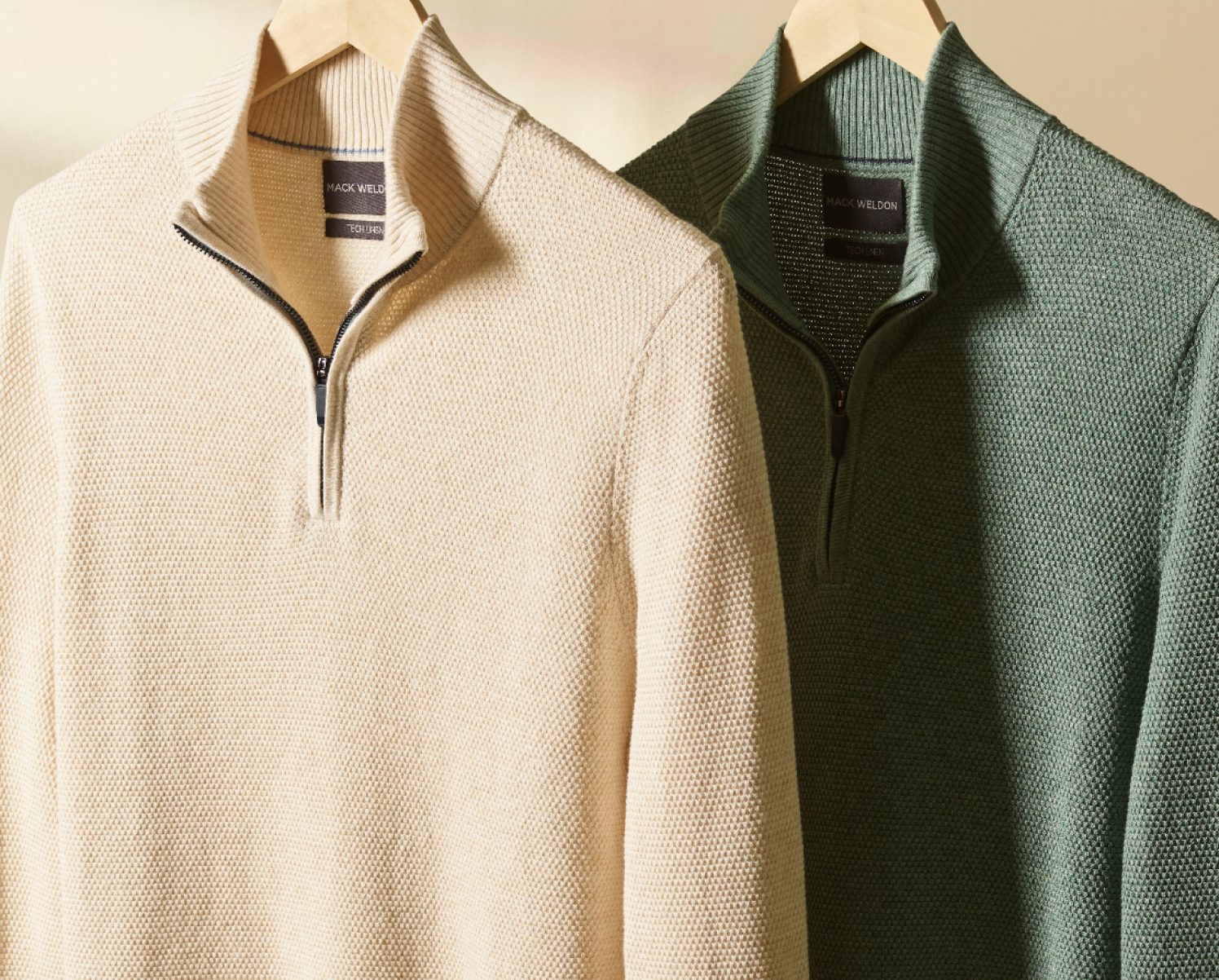 Image of two Tech Linen half zips, one cream and one green, hanging from hangers